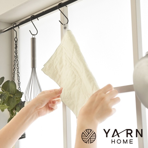 一件淺米色、絎縫紋路的廚房擦拭巾，由脫脂棉混紗製成，由雙手拿著掛在掛鉤上。