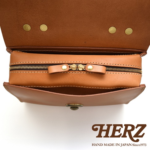 日本HERZ職人手作皮革多功能收納單肩包，近距離拍攝，展示了包包的拉鍊和翻蓋細節，包身為焦糖棕色，品牌標誌HERZ及產地字樣位於畫面下方