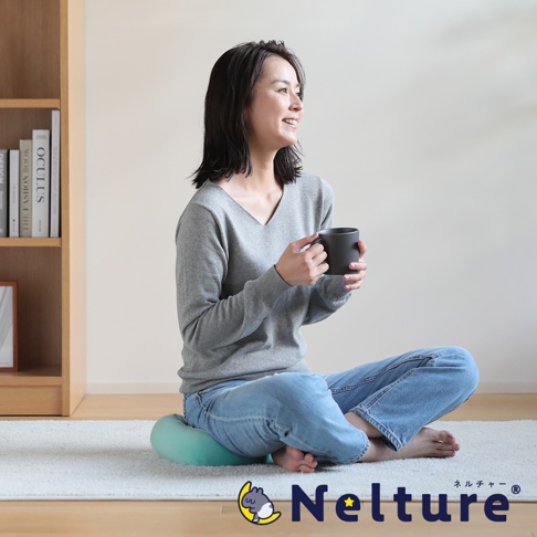 一名女性坐在土耳其藍色坐墊上，雙腿盤起，手裡拿著馬克杯，臉上帶著微笑看向窗外。