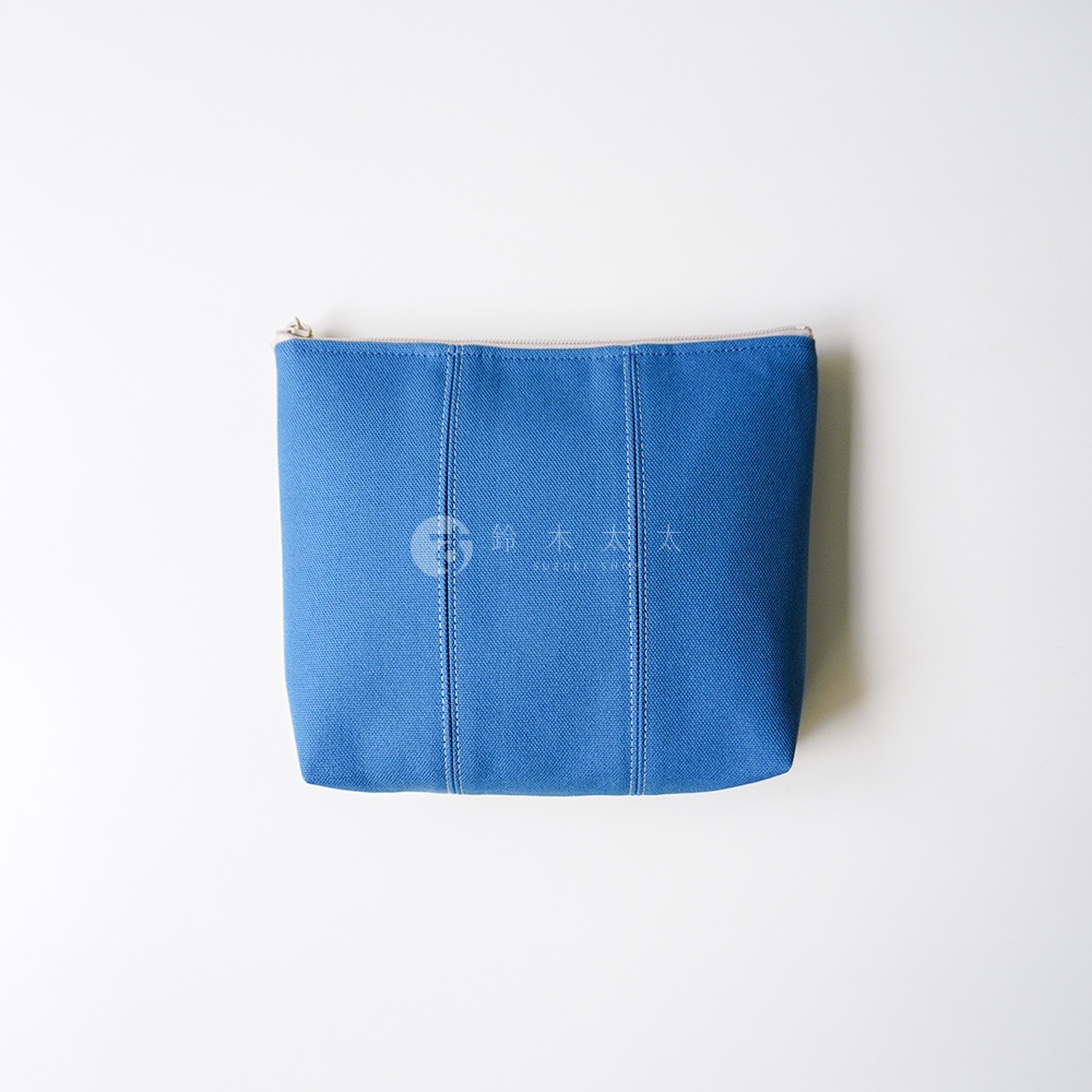 一個簡約的藍色帆布化妝收納包，表面帶有細緻的織紋質感，包身正面由垂直車縫線分隔為三個區塊，頂部設有拉鍊開口，整體造型洗練且具備良好的收納功能，中間印有淺灰色的鈴木太太品牌標誌。