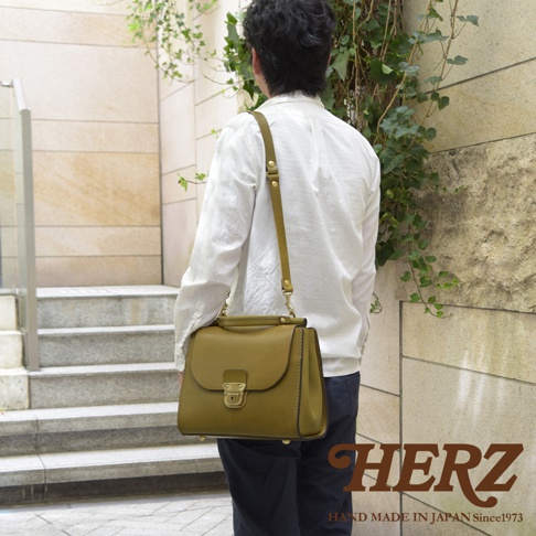 日本HERZ職人手作的橄欖綠皮革瓦風兩用單肩包，男性模特斜背於肩，包款的鎖扣與金屬細節清晰可見，背景是帶有植物的磚牆與階梯。