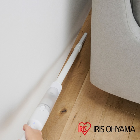 白色超輕量多合一手持無線吸塵器，長管連接扁平吸嘴，正在靠近淺灰色沙發與白色牆壁之間的縫隙，底部木質地板。