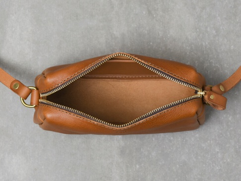 頂視角拍攝的棕色皮革拉鍊單肩包，拉鍊部分打開，露出內部。包身呈現自然的皮革紋理，搭配古銅色金屬拉鍊。