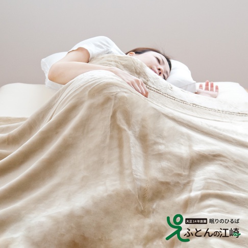 一名女子蓋著一條淺棕色、觸感柔軟的加厚毯子，毯子表面有細緻的紋理，展現其柔暖材質。