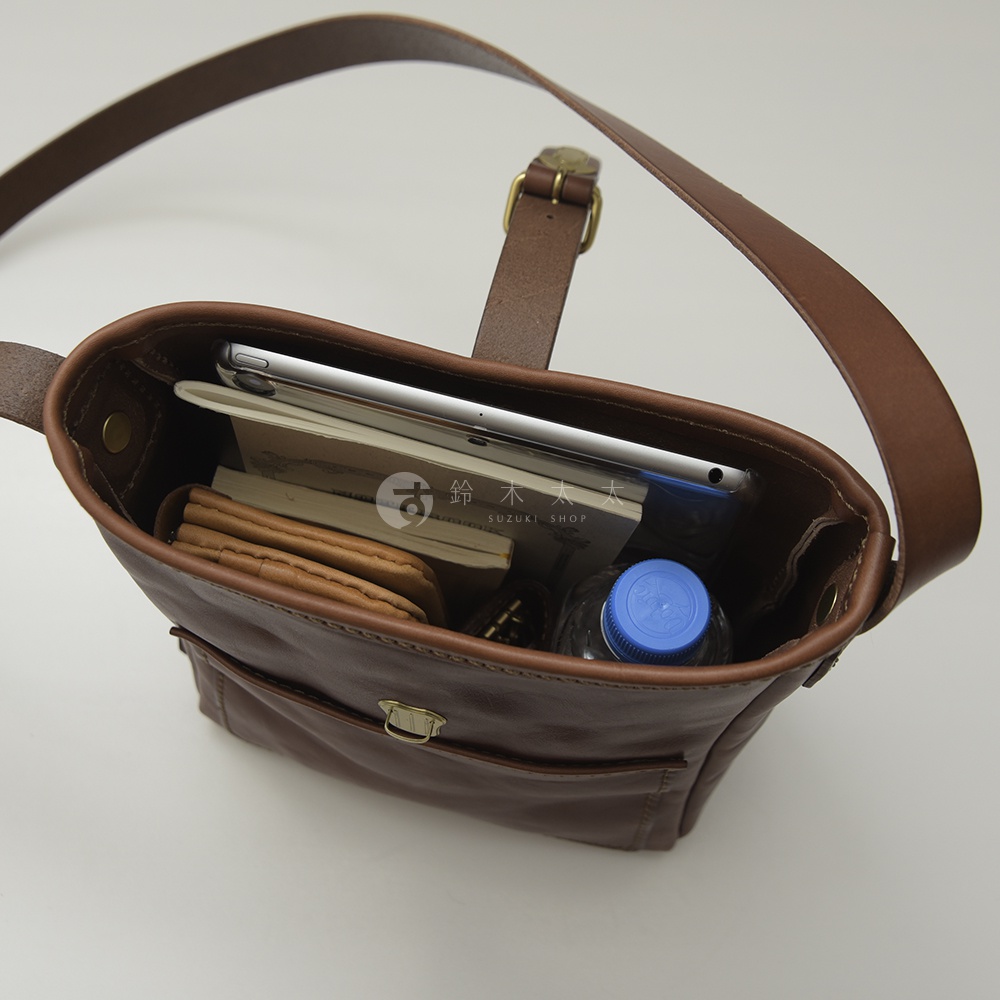 深棕色職人手作皮革輕便單肩側背包，內部裝有平板電腦、筆記本、皮夾和一瓶藍蓋礦泉水，展現其收納空間和材質細節。