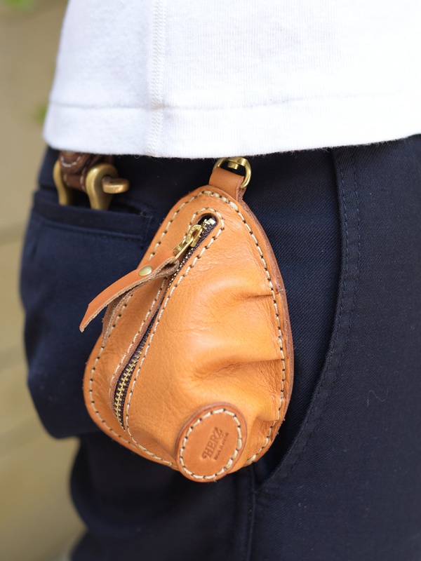 一個棕色皮革製成的餃子造型零錢包，帶有金色拉鍊和鑰匙圈，掛在深藍色褲子的腰帶環上。