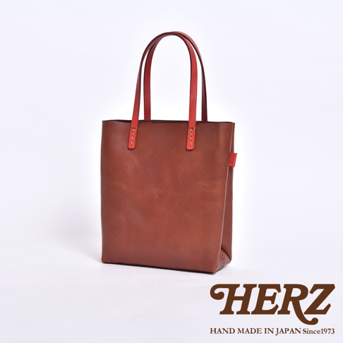 這是一款由日本品牌 HERZ 職人手工製作的直式軟皮托特包，呈現簡約的皮革質感。包身選用棕褐色皮革，搭配兩條醒目的紅色手把，側面飾有紅色的品牌織標，整體設計俐落且具備手作工藝感，背景為乾淨的純白底色。