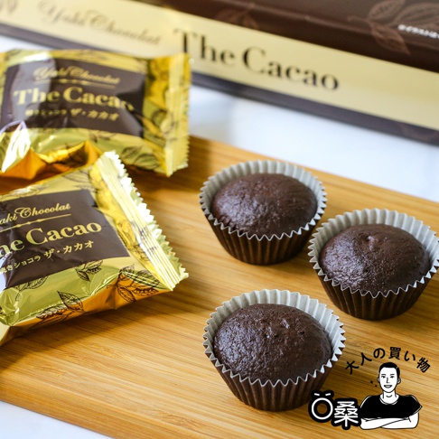三個獨立包裝的深棕色烤巧克力菓子，放在木質托盤上，兩旁是金黃色包裝的同一款巧克力，包裝印有「The Cacao」字樣。