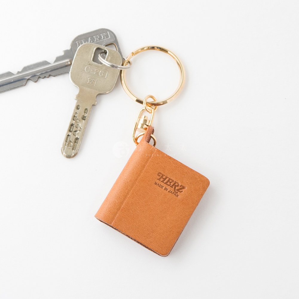 職人手作皮革書本造型鑰匙圈，呈現焦糖棕色，材質為真皮。鑰匙圈上印有「HERZ MADE IN JAPAN」字樣，搭配金色金屬配件及兩把鑰匙。
