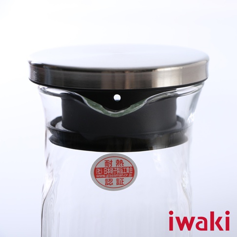 透明耐熱玻璃水壺，搭配銀色不鏽鋼蓋與黑色密封圈，壺身中央貼有紅色圓形認證標籤。
