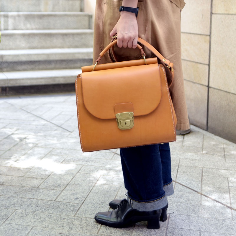 一款淺棕色皮革製的兩用單肩包，包身方形，帶有翻蓋和金色鎖扣，手柄圓潤，縫線清晰。