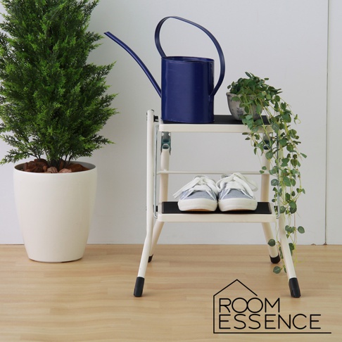 白色金屬折疊樓梯椅，兩層踏板，上面擺放著一對淺藍色帆布鞋，右側擺放著藍色灑水壺和一盆綠色藤蔓植物，左側有一盆綠色植物。