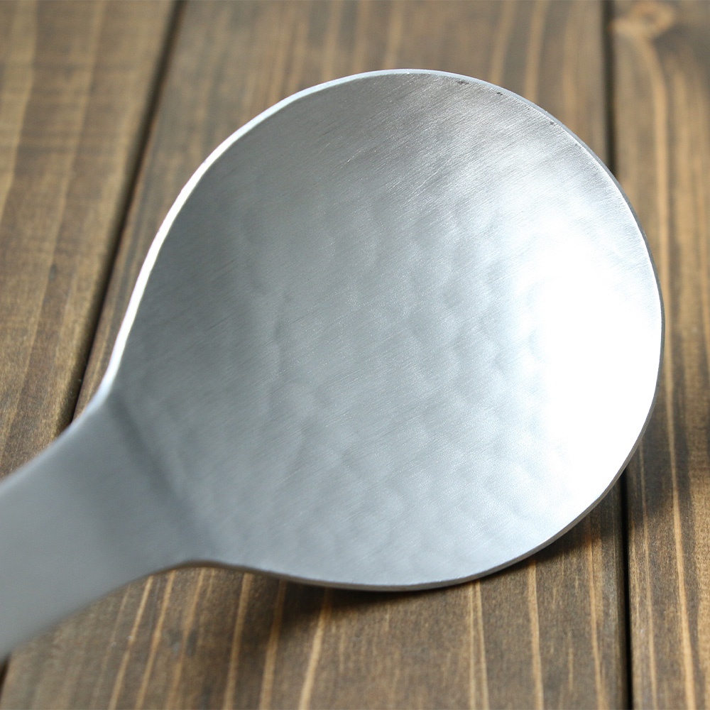 一個銀色的鋁製湯勺，勺面有錘目紋，勺柄連接處呈現弧形，放置在木紋背景上。
