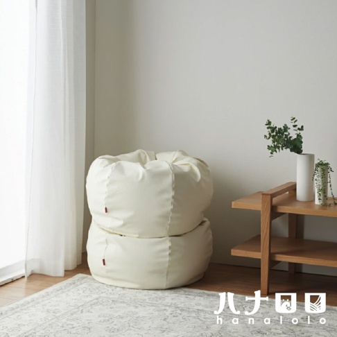 兩個堆疊的米白色皮革懶骨頭沙發，左側有白色窗簾，右側有木製邊桌，桌上有花瓶和植物。