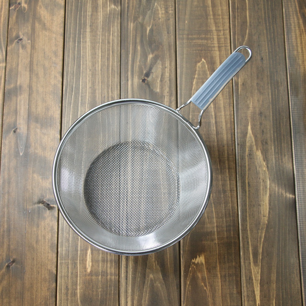 不鏽鋼圓形濾網，網孔細密，配有灰色矽膠防滑握柄，置於深色木紋桌面上。