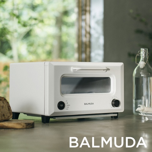 ReBaker 烤麵包機，白色，現代簡約設計，配有銀色把手和黑色旋鈕。前方有兩個控制旋鈕和一個模式選擇按鈕，以及一個透明玻璃烤箱門，可見內部烤架。產品放置在淺灰色檯面上，背景為模糊的綠色植被和一個玻璃瓶。