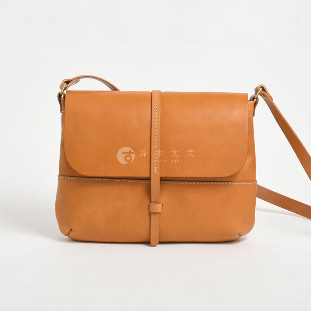 一個以經典棕色皮革製成的斜挎包，包蓋上有細緻的縫線裝飾，並透過細皮帶固定，包款中間有一個日文標誌和文字。