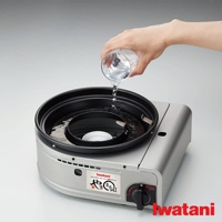 銀色金屬外殼的 Iwatani 無煙燒烤爐，上方黑色烤盤正在注入清水，烤爐側面有品牌標誌和控制旋鈕。