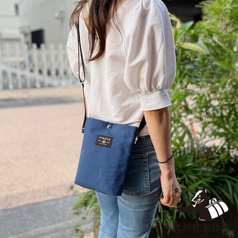 一位女性身穿白色寬袖上衣和牛仔褲，斜挎一個藍色帆布小方包，包包上有一個黑色標籤，上面印有日文文字和圖案。