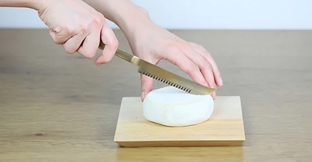 一隻手正用一把金屬質地的起司刀，切著放在木質砧板上的白色圓形起司。起司刀刀刃帶有鋸齒狀。