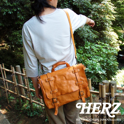 職人手作水洗加工皮革復古郵差包，以溫暖的橘棕色皮革製成，配有可調節的長肩帶、頂部提把，以及兩排帶有黃銅釦的皮帶裝飾，呈現經典郵差包造型。