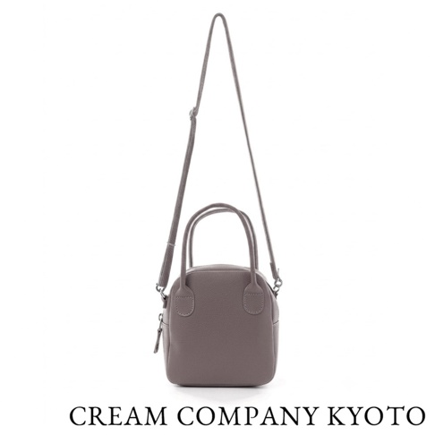 CREAM COMPANY 京都出品的藕灰色皮革貝殼包，設計簡約精緻，配有雙提把與可拆式細肩背帶，包身呈現溫潤的皮革紋理，底部標註品牌名稱，適合手提或肩背使用。