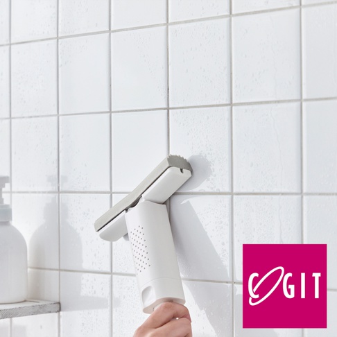 白色推拉瀝水刮刀，帶有灰色橡膠刮片，手柄上有圓形透氣孔，正被一隻手握住，用於清潔佈滿水珠的白色瓷磚牆面。