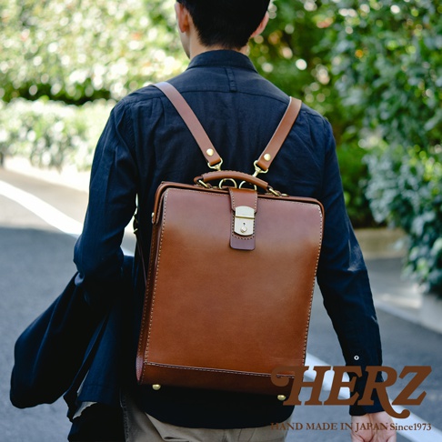 一名男子身背棕色皮革製杜勒斯商務後背包，包款為方形設計，有金屬釦環與提把，肩帶為皮革材質。