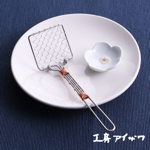 銀色不鏽鋼製成的方形豆腐杓，帶有細網狀勺面，銅色線條纏繞的握柄。旁邊有一個白色小碟，碟上有花朵圖案。