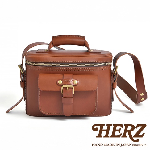HERZ 經典款棕色皮革相機包，配有頂部提把、可調式肩帶和前置翻蓋口袋，口袋上飾有復古皮帶扣和 HERZ 品牌標誌，下方印有「HAND MADE IN JAPAN Since1973」字樣