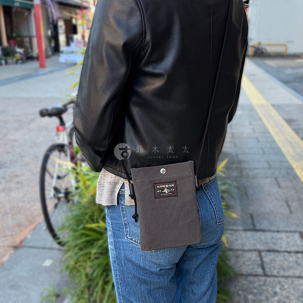 一位身穿黑色皮夾克和牛仔褲的人，斜挎著一個犬印鞄製作所的灰色帆布手機包。