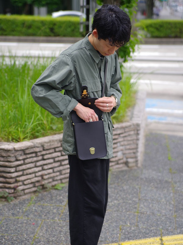 一位男士身穿軍綠色襯衫，斜背著一個黑色皮革單肩包，包款有著經典的皮扣設計，整體呈現簡約風格。