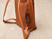 職人手作皮革後背包，採簡約褶皺風設計，主體為溫潤的棕色皮革，配有黃銅拉鍊及可調節的皮革肩帶。包側有一個拉鍊口袋，露出一個同色系的皮革零錢包。