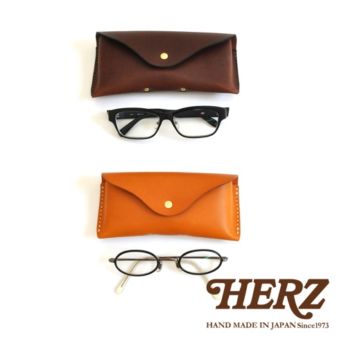 兩個皮革眼鏡收納盒，上方為棕色，下方為橘色，分別搭配一副黑色方框眼鏡和一副圓框眼鏡。右下角有HERZ品牌標誌。