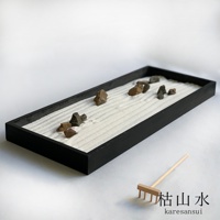 黑色長方形木製沙盤，內有細膩的白色沙子，耙出波浪紋理，散布著幾塊深淺不一的灰色、棕色石塊。右側有一迷你木製耙子，沙盤旁有「枯山水」中日文標示。