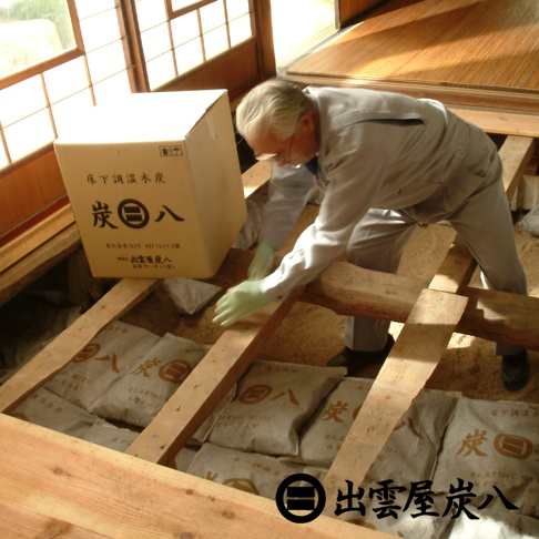 一群工人正在地板下方放置多個裝有木炭的白色袋子，其中一個大紙箱上印有「炭八」字樣。