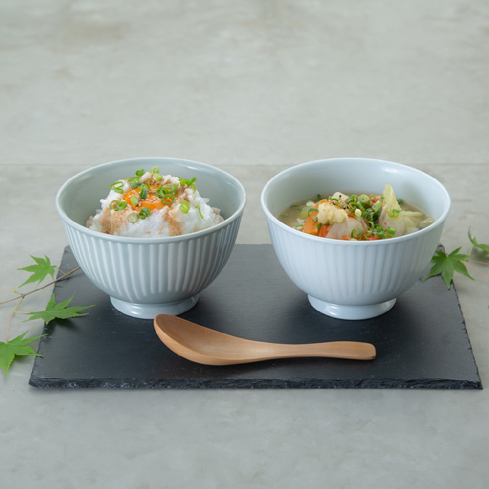 兩個淺藍色帶有細緻垂直紋路的陶瓷飯碗，分別盛裝著飯和湯。飯碗旁有一支木製湯匙，背景為淺灰色桌面，左側點綴著綠色楓葉。