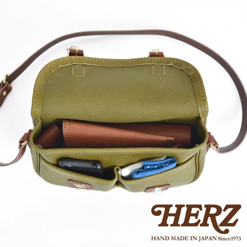 橄欖綠皮革傳統釣魚包，內部有深棕色皮革內袋，前袋放置手機及藍色圖案織品，搭配棕色皮革肩帶，包款為HERZ品牌。