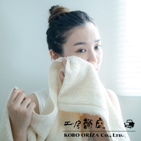 一位女性正在使用一條淺米色、超細纖維毛巾擦拭臉部。毛巾質地蓬鬆，邊緣有精緻的縫線。