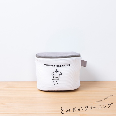 白色網狀洗衣袋，附有灰色網狀提蓋，袋身印有「TOMIOKA CLEANING」字樣及一個衣架吊著一件衣服，下方有水滴圖案。