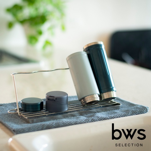 不鏽鋼折疊杯壺瀝水架上，放置著兩個不同顏色的保溫杯，分別為淺灰色和深藍色。瀝水架下方有兩個深灰色的小蓋子，整個瀝水架置於一塊藍灰色毛巾上。