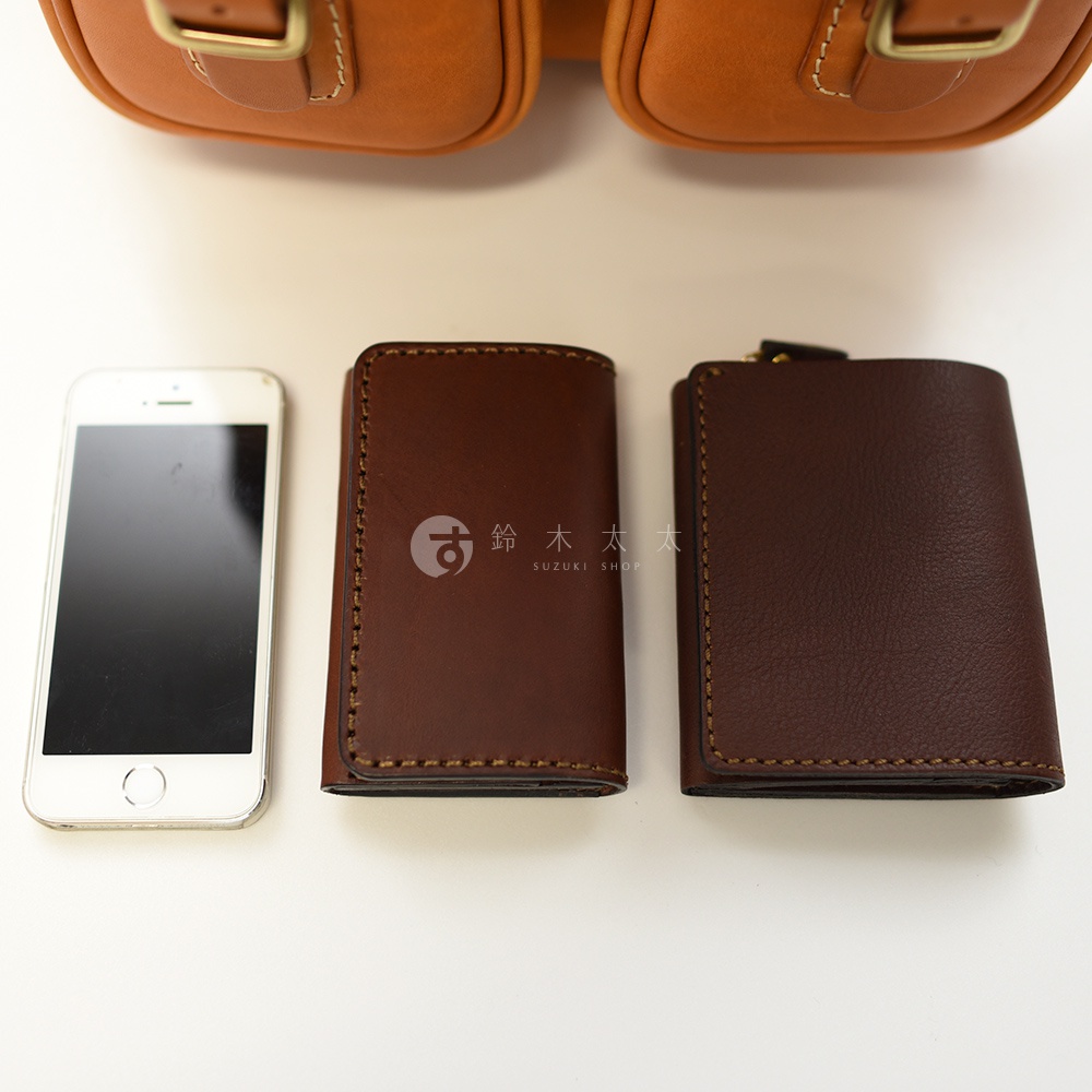 三種深棕色皮革製品：一個iPhone手機、一個較小的方形錢包，以及一個較大的拉鍊錢包，置於淺色背景上。