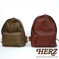 兩個職人手作皮革後背包，一個為橄欖綠，另一個為紅棕色。兩者皆為休閒風格，採用經典設計，配有雙向拉鍊主隔層、前方拉鍊口袋，以及菱形皮革標誌。