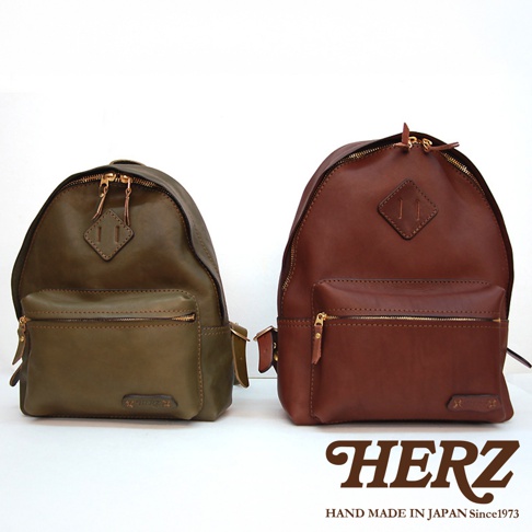 兩個職人手作皮革後背包，一個為橄欖綠，另一個為紅棕色。兩者皆為休閒風格，採用經典設計，配有雙向拉鍊主隔層、前方拉鍊口袋，以及菱形皮革標誌。