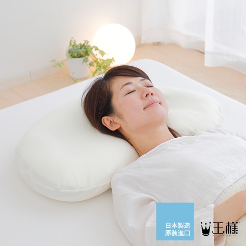 一位女性仰臥在白色床鋪上，頭部枕著一個白色、柔軟的雲狀枕頭。枕頭呈彎月形，表面光滑，材質看似布料。背景有植物與圓形燈飾。