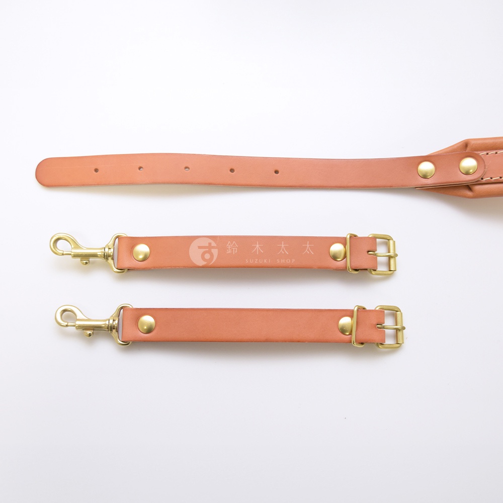 職人手作皮革加厚減壓肩背帶，展示三段式設計，包含一條較長的肩帶和兩條較短的連接帶。整體為淺棕色皮革材質，搭配黃銅色金屬扣環與鉚釘，其中一條連接帶上印有「鈴木太太 SUZUKI SHOP」的品牌標誌。