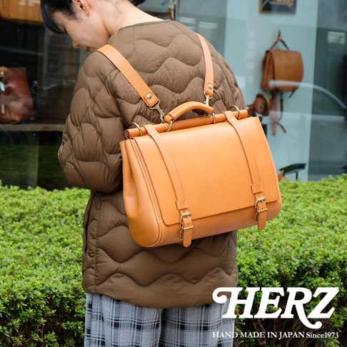 一位女性背著一個日本HERZ品牌的手工皮革瓦風側背包，包包為橘褐色，有著金屬扣環和提把，搭配她身上褐色的菱格紋外套和格紋長褲。