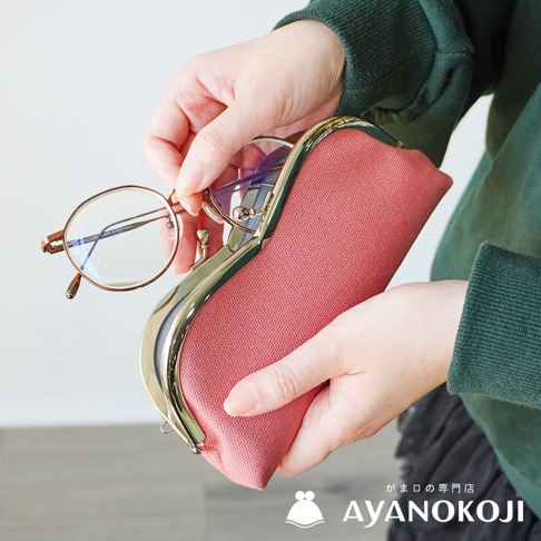 一雙手正將金屬框眼鏡放入桃紅色帆布質地的口金眼鏡收納盒中，收納盒旁有「がま口の専門店 AYANOKOJI」的品牌標示