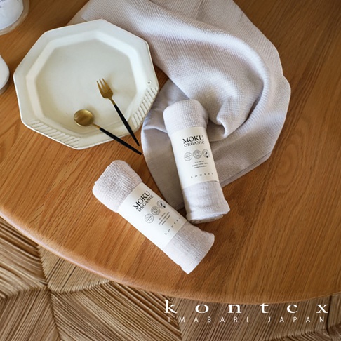 兩條 MOKU ORGANIC 有機棉長毛巾，一條為淺米色，另一條為淺灰色，均捲成長筒狀，標籤清晰可見。旁邊擺放著一個八角形白色盤子，盤內有一支金色叉子和一把金色湯匙，盤子下方有一塊淺灰色的長毛巾。