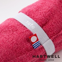 粉紅色毛巾捲起，繫有白色束帶，束帶上有 soto 字樣，一旁另有紅色小標籤，下方印有 HARTWELL MABARI JAPAN 字樣